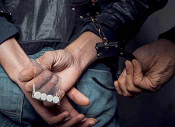 Person in handcuffs holding drugs, symbolizing drug-related arrests, controlled substance offenses, and law enforcement. - Law Office Of Greg Webb